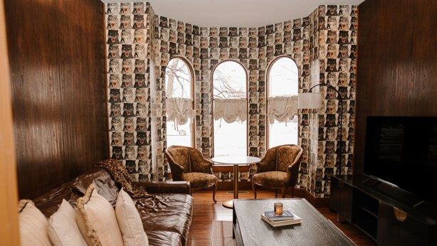 A cozy living room with a brown leather sofa, two armchairs, a coffee table, and a TV. The room features patterned wallpaper, large windows with curtains, and a wooden floor. - Oregon District Oasis - The Best of Downtown (Dayton)
