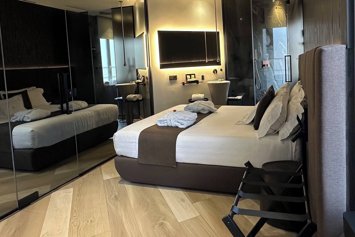 A modern hotel room with a large bed, a seating area, a vanity with a mirror, and a large window.
