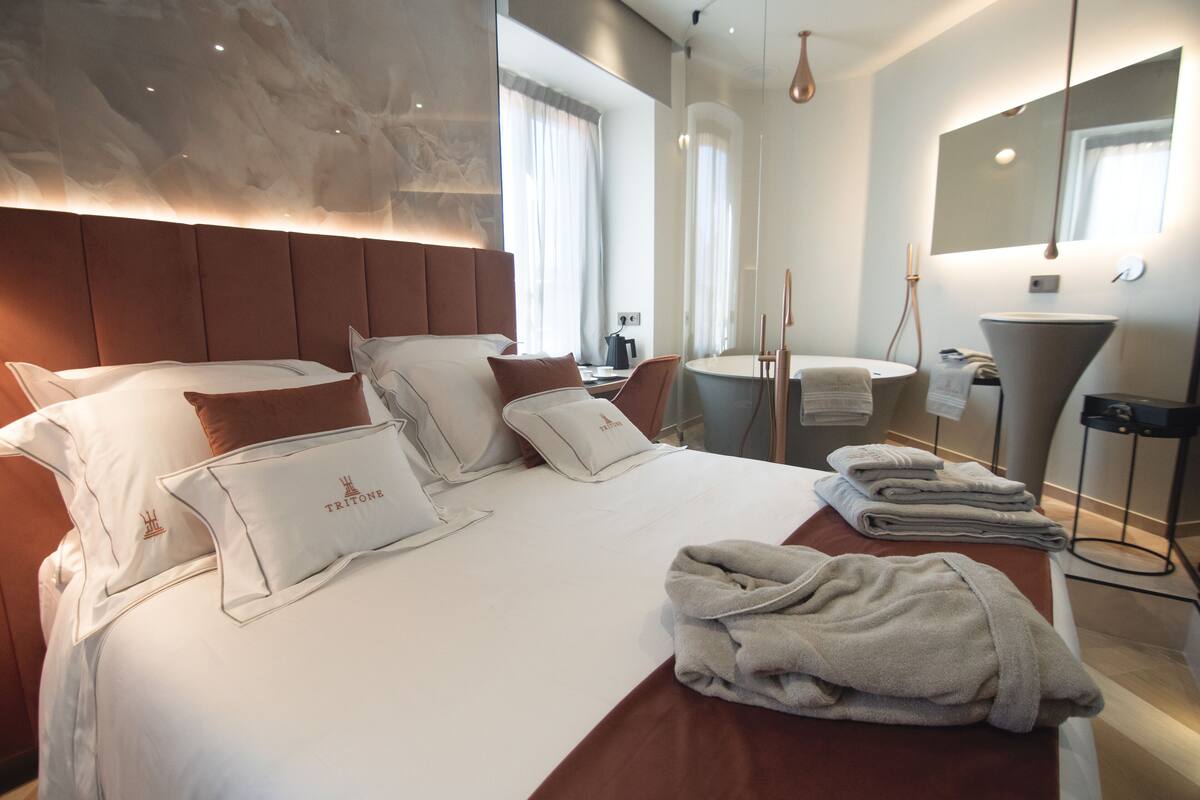 A modern hotel room with a large bed, multiple pillows, a bedside table with a lamp, a round mirror, and a bathroom area with a sink and towel rack.