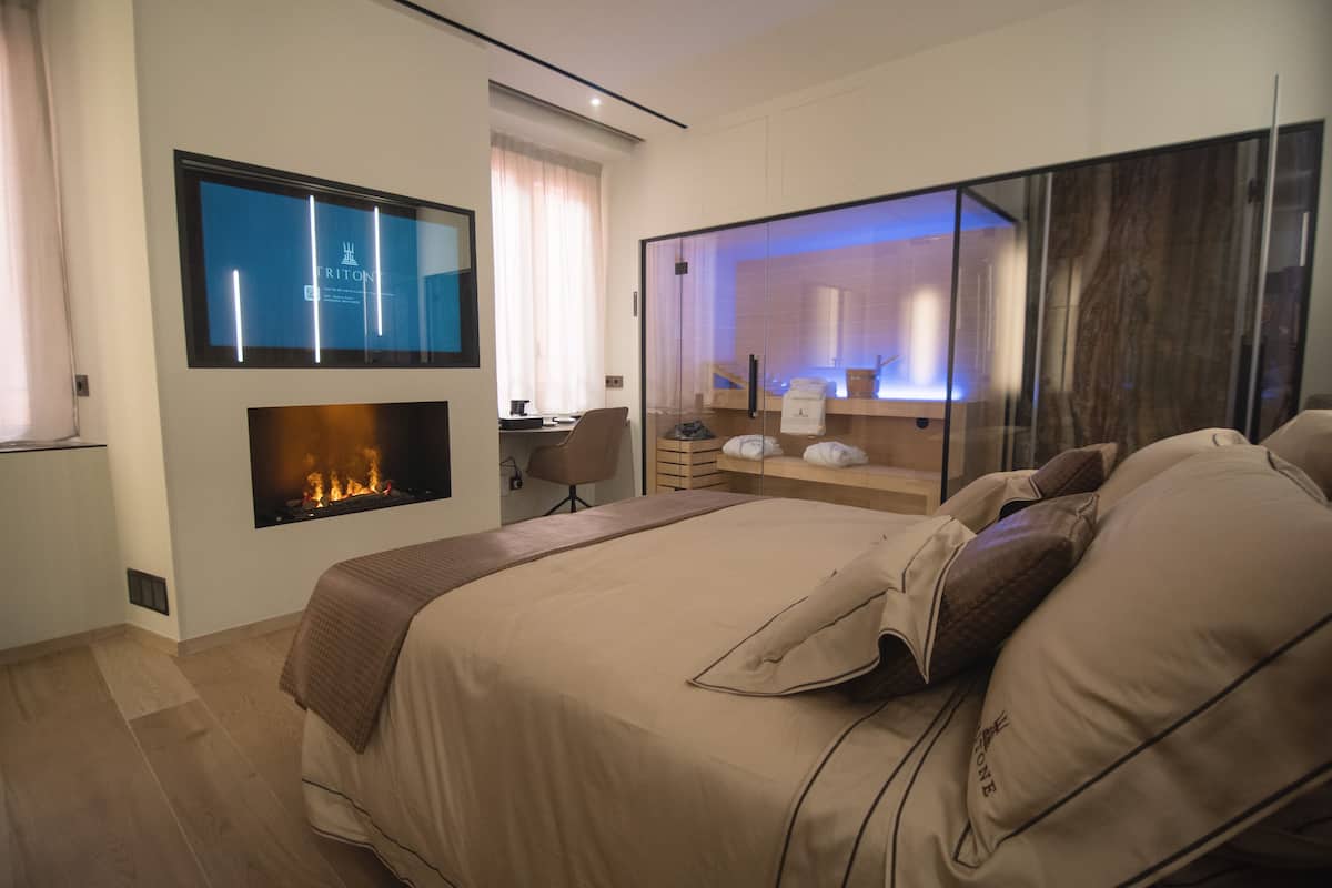 A modern hotel room with a large bed, a fireplace, a desk, and a glass partition.