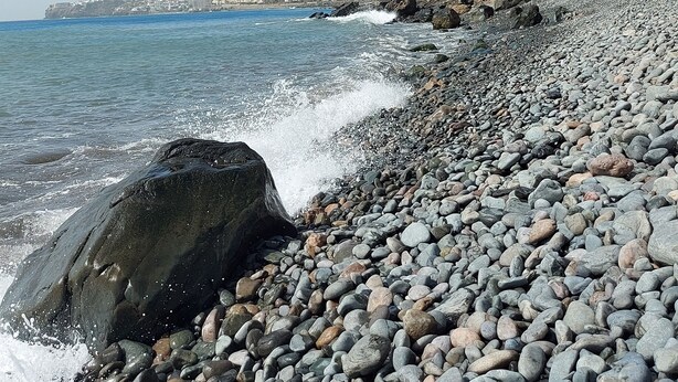 A rocky beach with waves crashing onto the shore, a stone staircase leading up to a building with palm trees, and a clear blue sky.