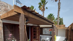 A patio area with a wooden pergola, stone walls, a white tiled floor, a table with chairs, and potted plants.
