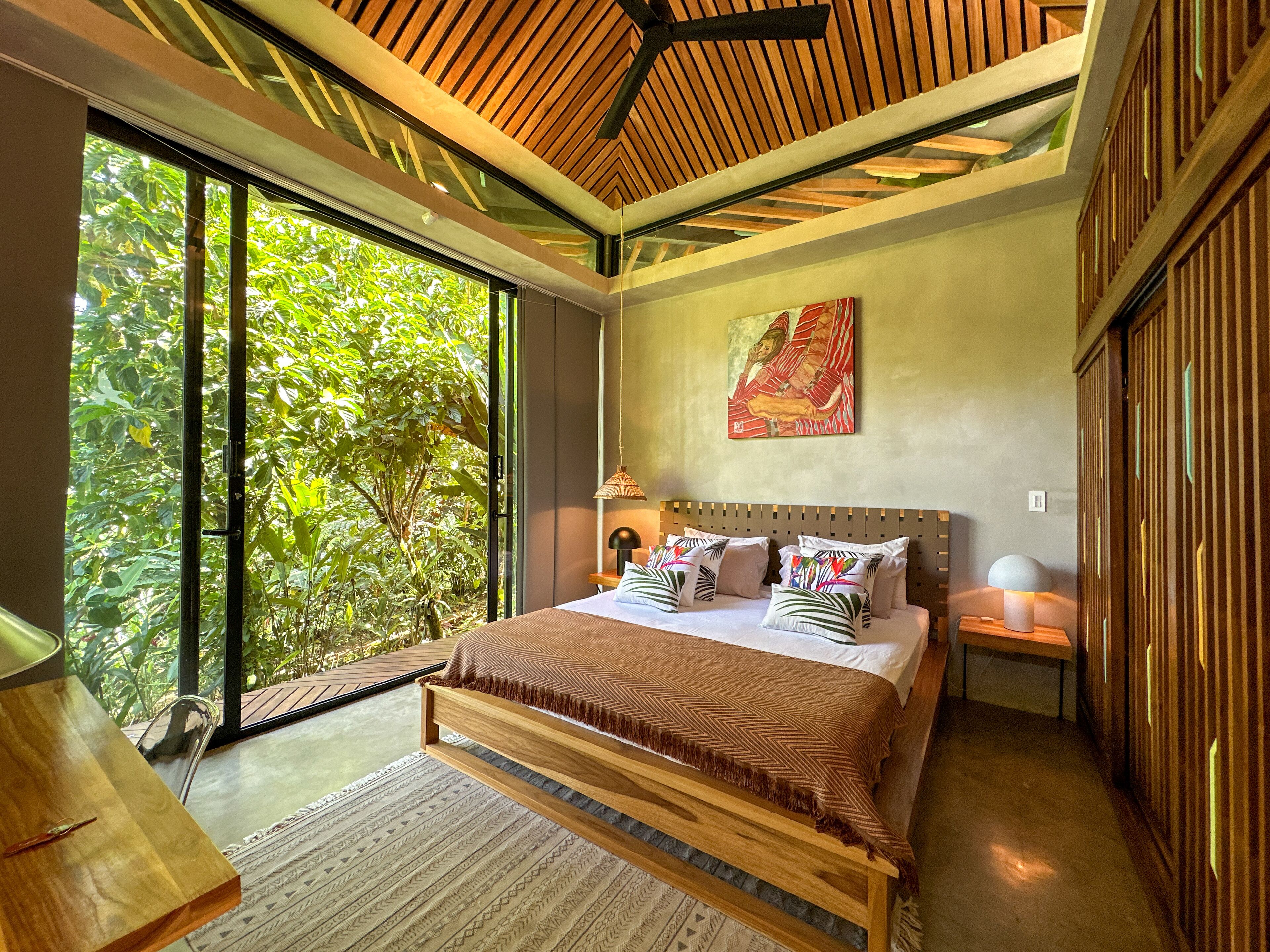 Ein modernes Schlafzimmer mit einem großen Bett, einem Holz-Kopfteil und Blick auf üppiges Grün durch raumhohe Fenster.