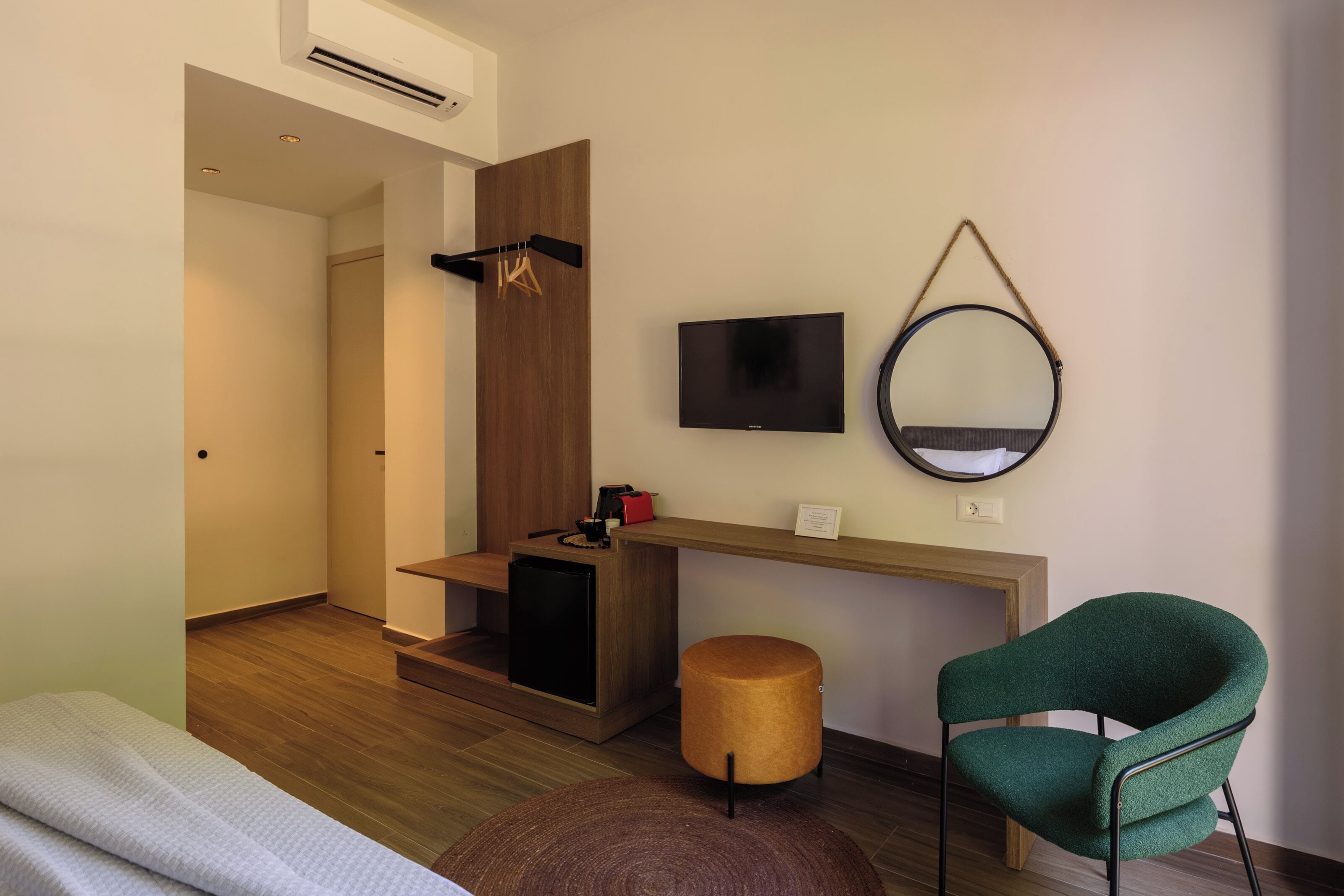 A modern hotel room with a wooden floor, a bed with a white cover, a small round ottoman, a green chair, a TV, a mirror, and a wooden shelf with a red bag and a black object.