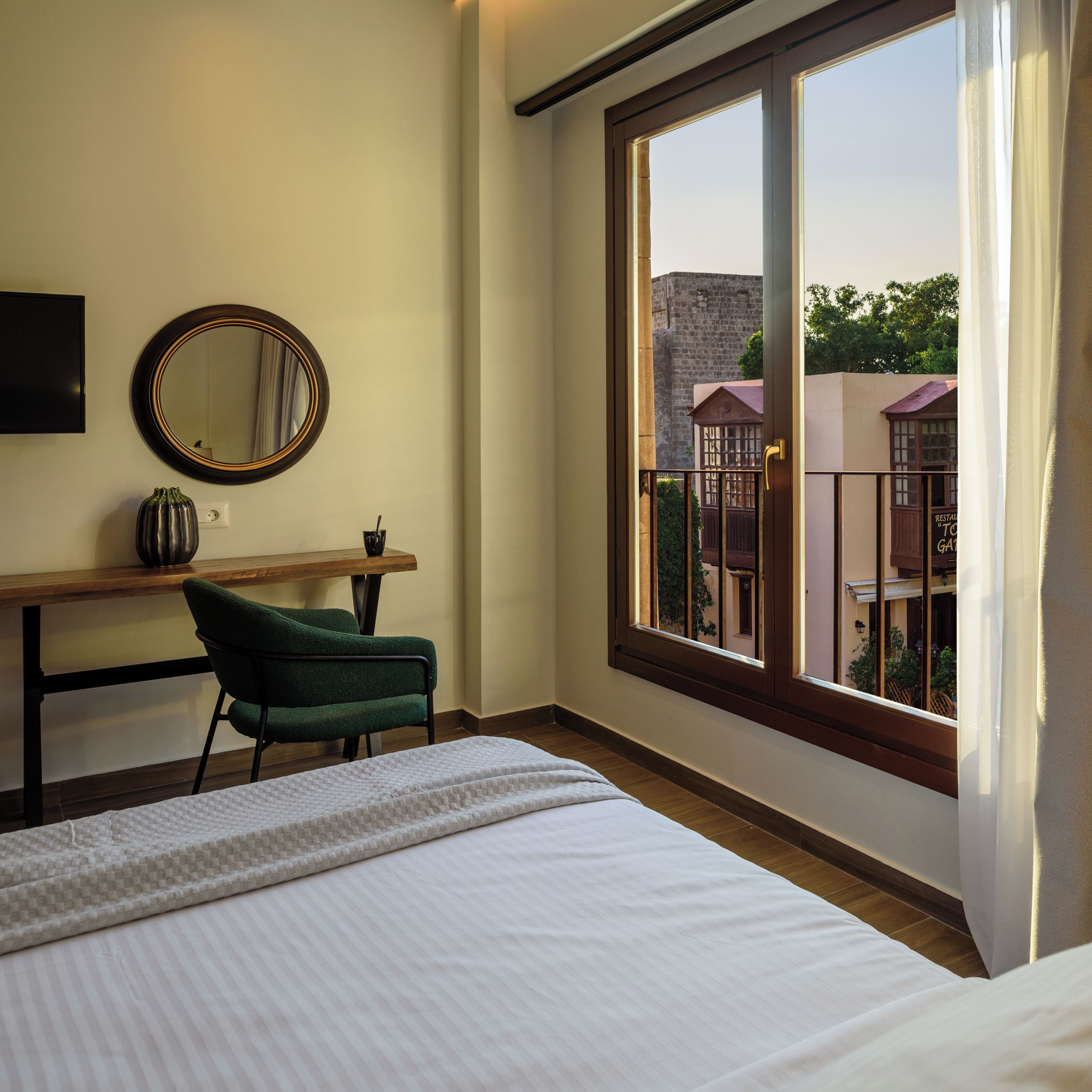 Una habitación de hotel con un ventanal grande, una cama, un escritorio, una silla y un espejo.