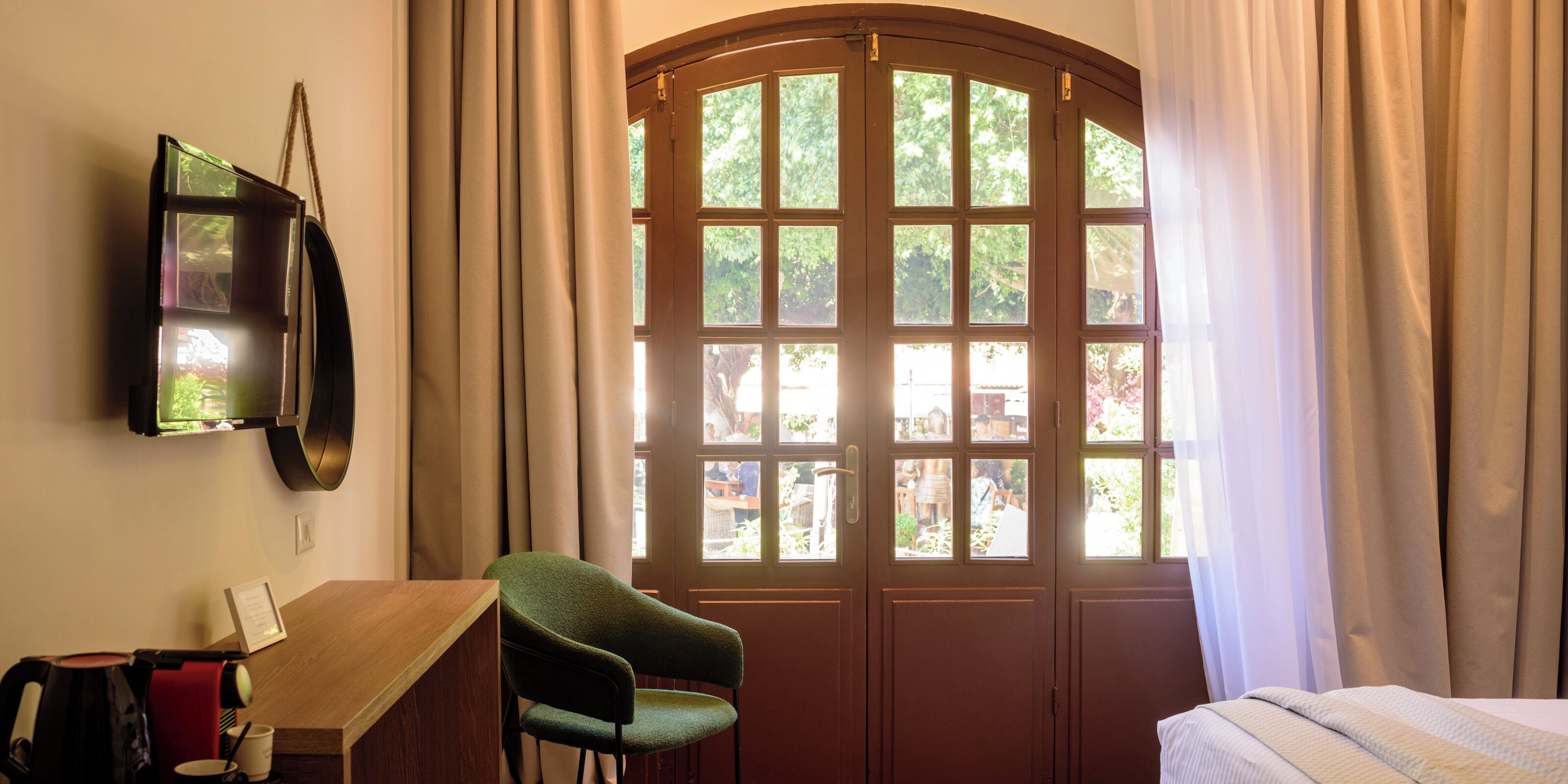 A room with a wooden desk, a green chair, a bed, and a large window with curtains.