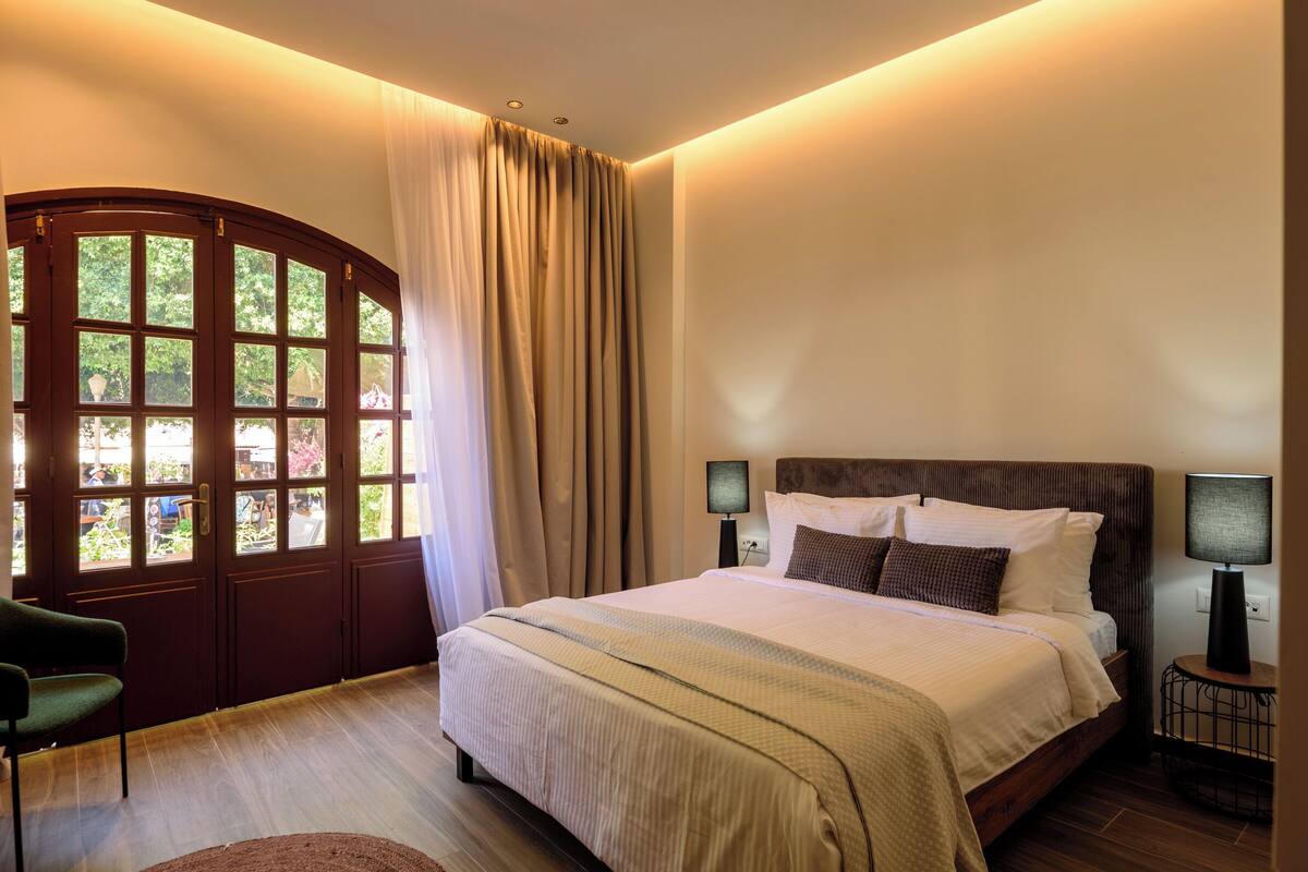 A bedroom with a bed, two bedside lamps, a chair, and a large window with curtains.