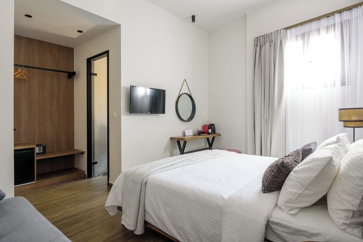 A modern hotel room with a bed, a grey sofa, a wooden wardrobe, a TV, and a bedside table with a lamp.