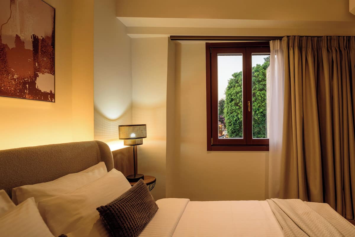 A hotel room with a bed, a nightstand with a lamp, a window with curtains, and a cityscape painting on the wall.