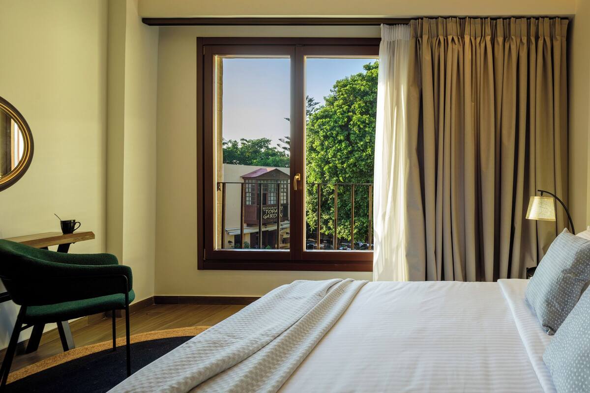 A hotel room with a large bed, a desk with a chair, a window with curtains, and a view of the outside.