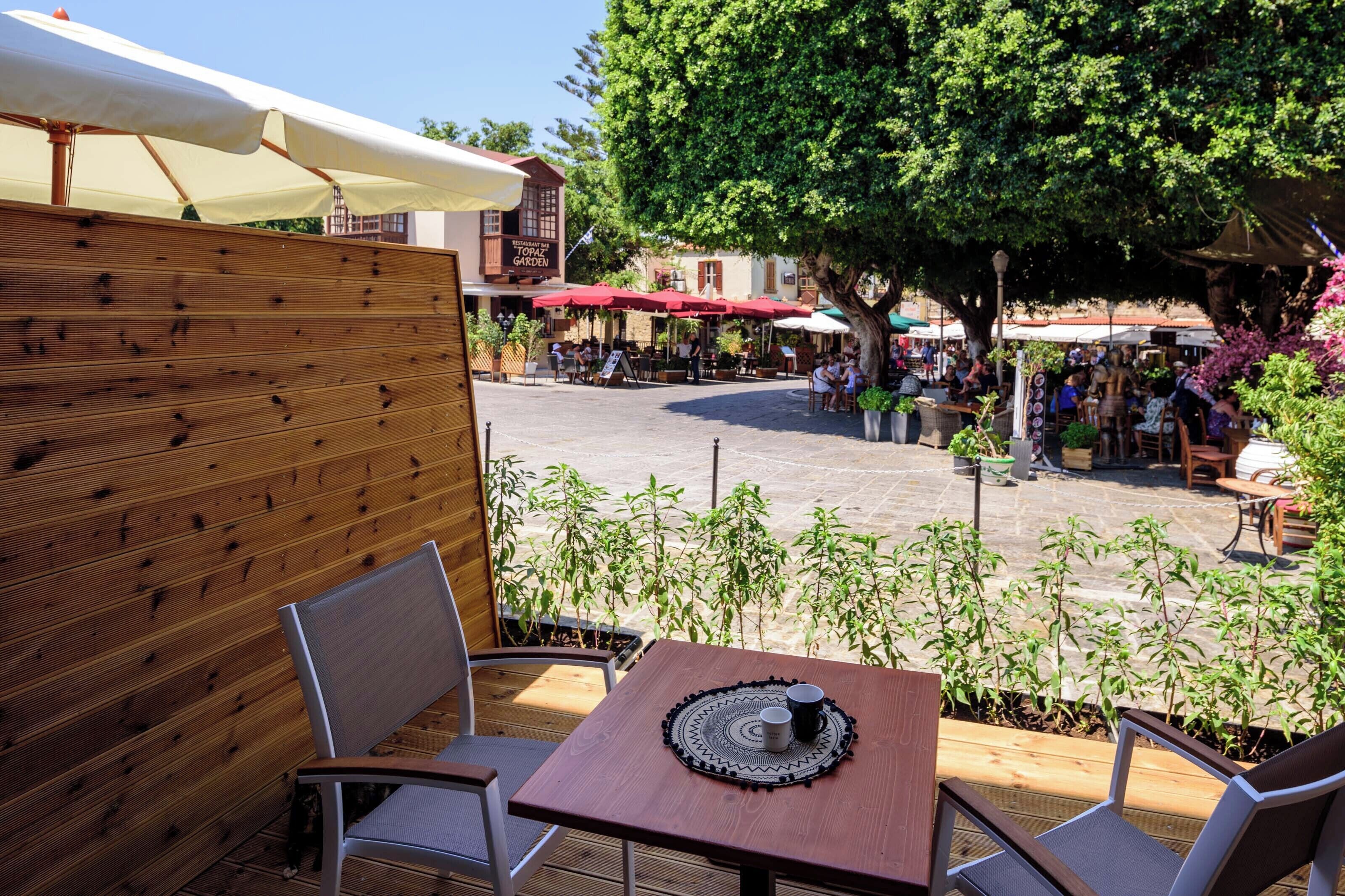Un espacio al aire libre con mesas y sillas de madera, rodeado de plantas en macetas y un gran árbol.