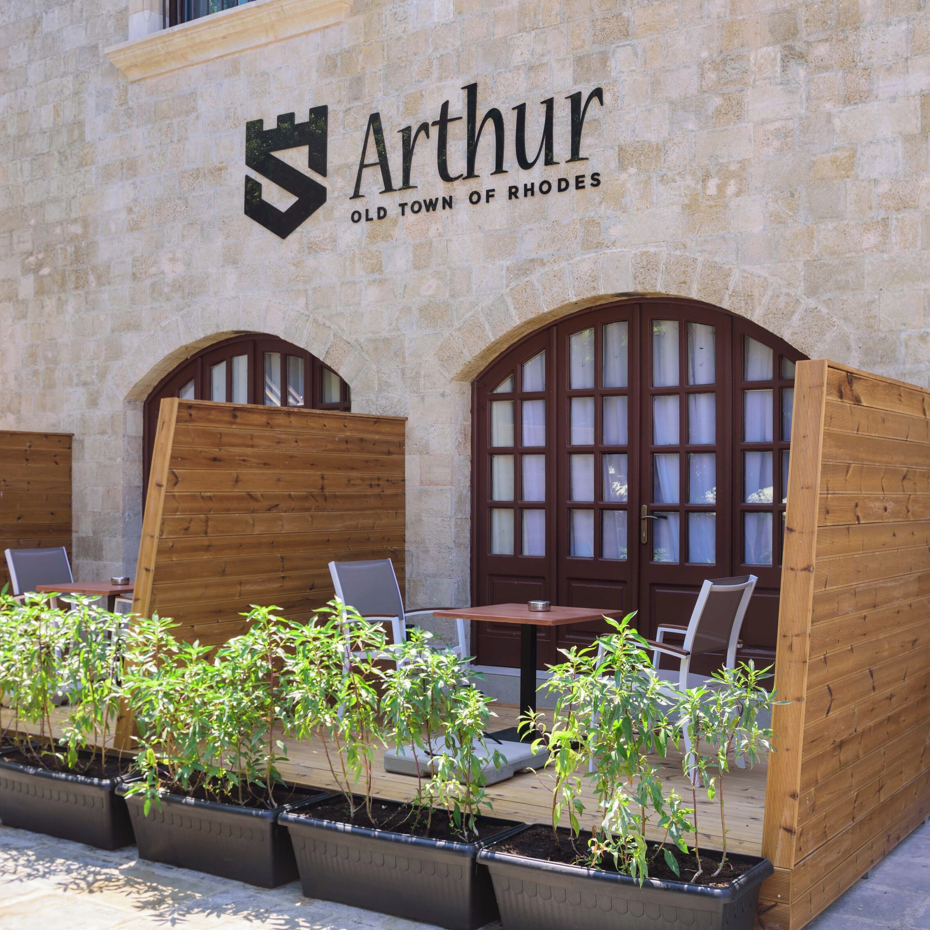 An outdoor seating area with wooden tables and chairs, potted plants, and a stone wall with the Arthur Old Town of Rhodes logo.