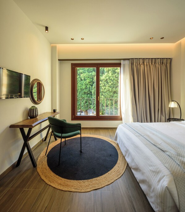 A bedroom with a bed, a desk with a chair, a TV, a window with curtains, and a lamp. - Arthur Suites (Rhodes)
