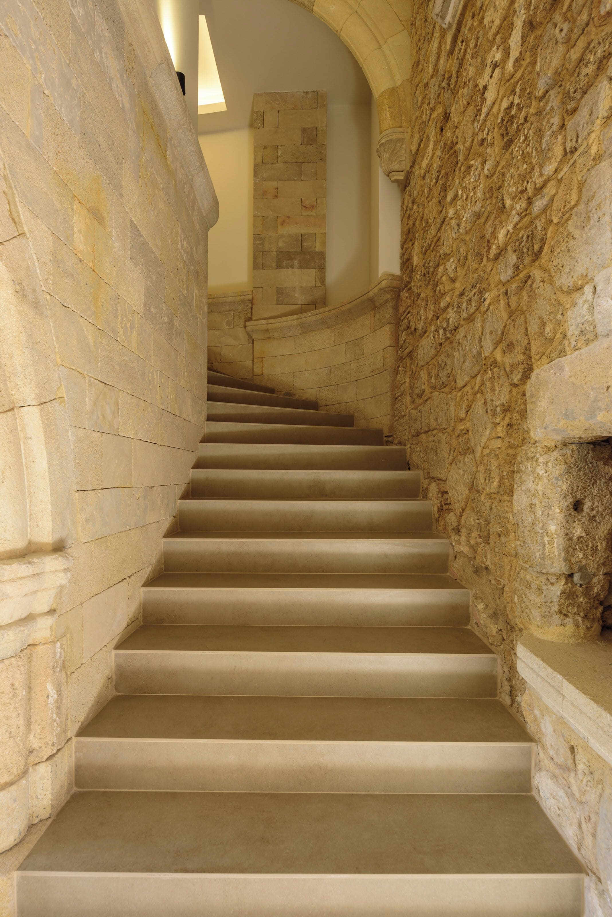 Un escalier en pierre, avec un mur en pierre d’un côté et un mur recouvert de carreaux de l’autre, mène à un niveau supérieur.