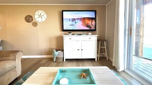 A living room with a TV mounted on the wall, a white cabinet, a wooden floor, and a window with a view. - Spacious 3-bedroom condo steps away from every thing in Ocean City! (Ocean City)