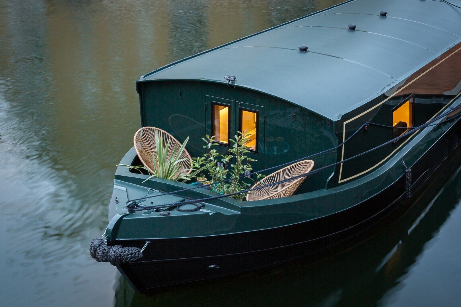 Une péniche à coque vert foncé, dotée de deux fenêtres éclairées d’une lumière chaleureuse et de chaises en osier sur le pont.