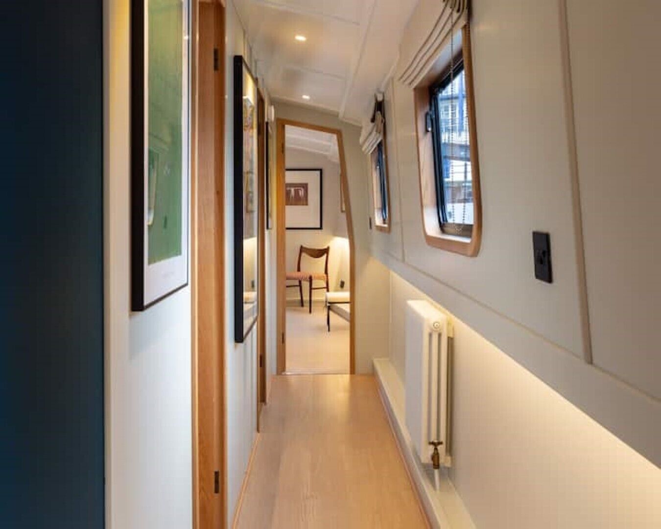 A narrow hallway with wooden flooring, white walls, and a radiator. There are framed pictures on the walls and a chair in the distance.
