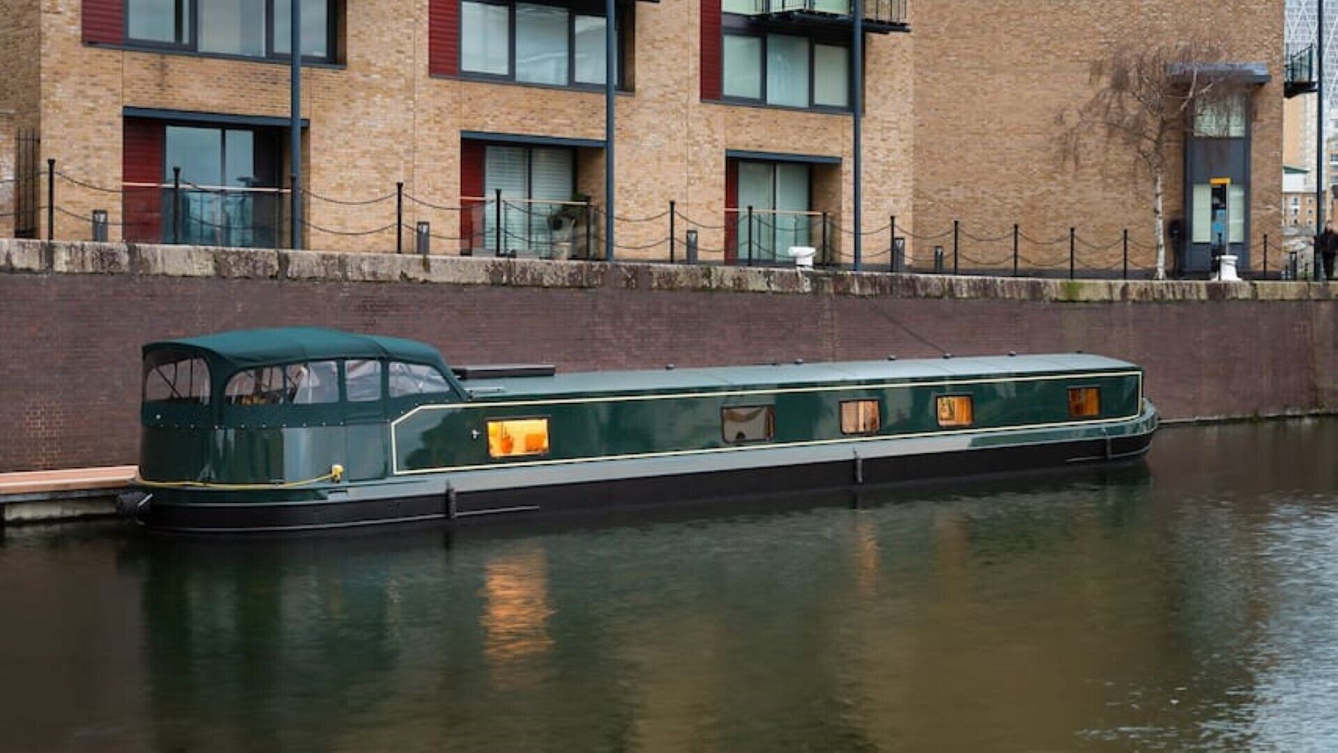 Un barco tipo narrrowboat atracado en un canal, con un edificio de ladrillo al fondo.