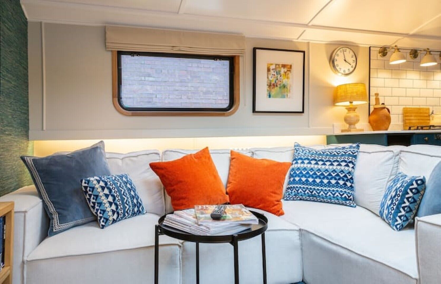 Un salon moderne avec un canapé blanc, des coussins aux motifs orange et bleu, une petite table ronde avec des magazines, une fenêtre donnant sur un mur de briques et une horloge au mur.