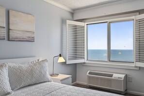 A bedroom with a bed, a nightstand, a lamp, a window with shutters, and a wall-mounted air conditioner. - Panoramic Ocean Views | Modern Kitchen|Heated Pool 1304 (Virginia Beach)