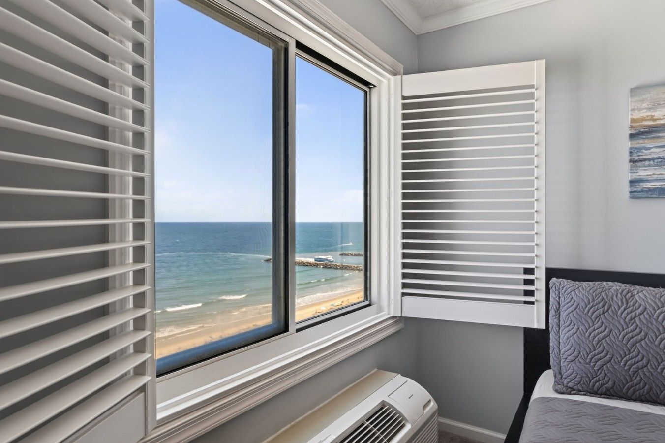 Une chambre dotée d’une grande fenêtre donnant sur une plage, d’un lit agrémenté d’un coussin à motifs et d’un climatiseur fixé au mur.