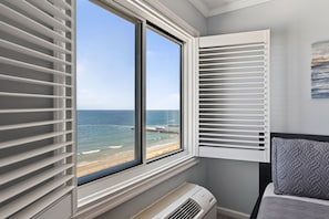 A room with a large window overlooking a beach, a bed with a patterned pillow, and a wall-mounted air conditioner.