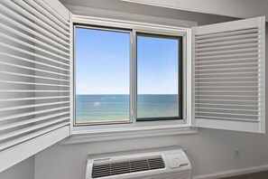 Une pièce avec une fenêtre donnant sur l’océan, un climatiseur blanc placé juste en dessous, et des stores blancs à la fenêtre.