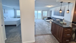 A spacious living area with a large window, a kitchen with dark wooden cabinets, and a balcony with a view. - Panoramic Ocean Views | Modern Kitchen|Heated Pool 1304 (Virginia Beach)