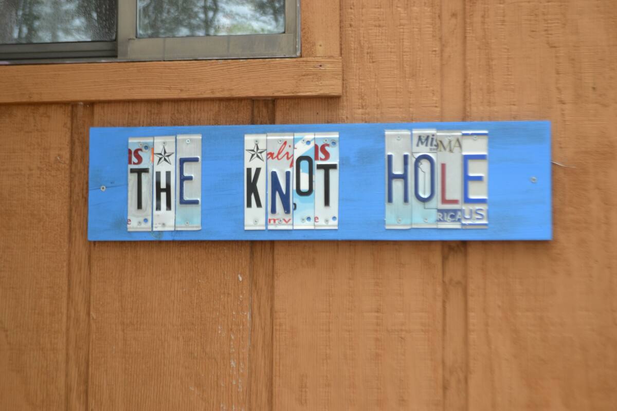 Eine Holzwand mit einem blauen Schild, auf dem Nummernschilder den Schriftzug "THE KNOT IS A HOLE" bilden.