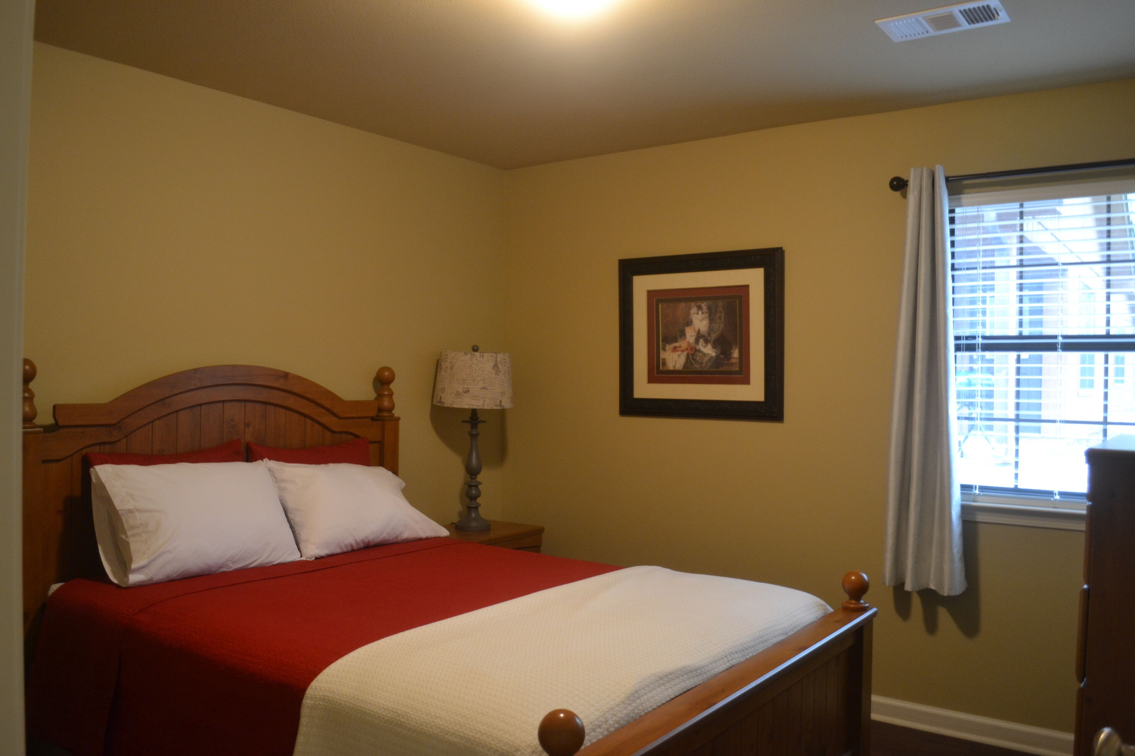 A bedroom with a wooden bed, a red and white bedspread, a nightstand with a lamp, a framed picture on the wall, and a window with blinds.
