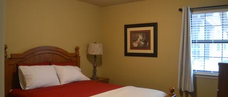 A bedroom with a wooden bed, a red and white bedspread, a nightstand with a lamp, a framed picture on the wall, and a window with blinds.