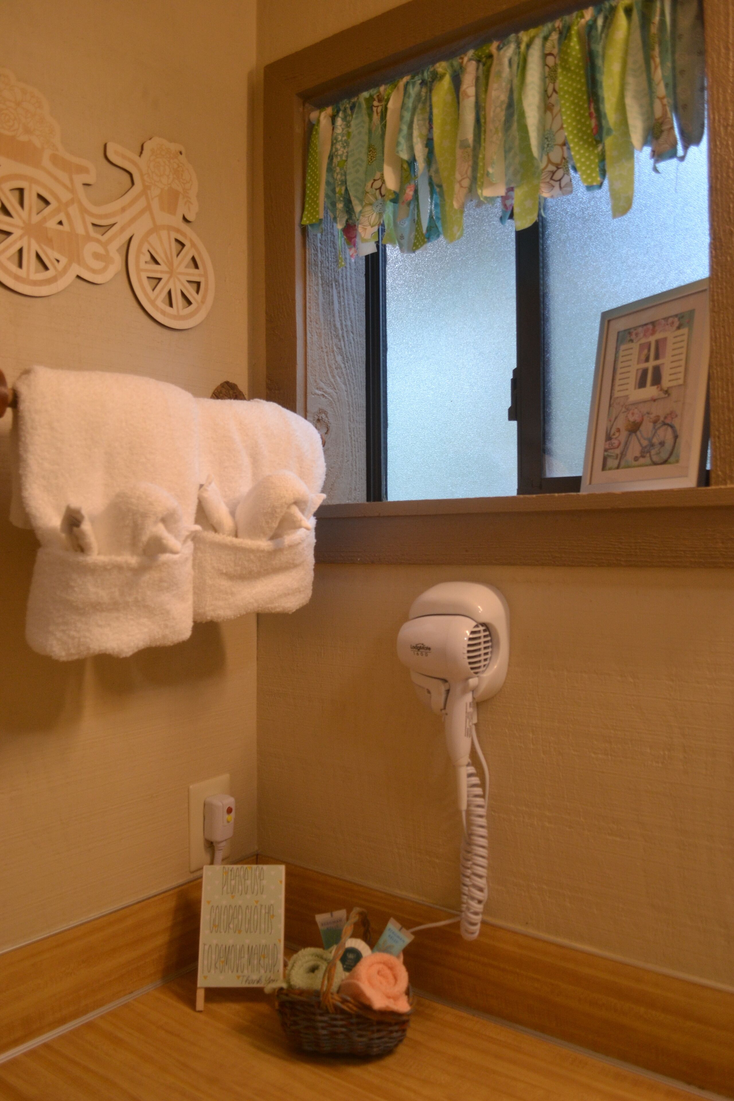 A bathroom with a towel rack, a hairdryer, a basket with toiletries, and a window with a curtain.