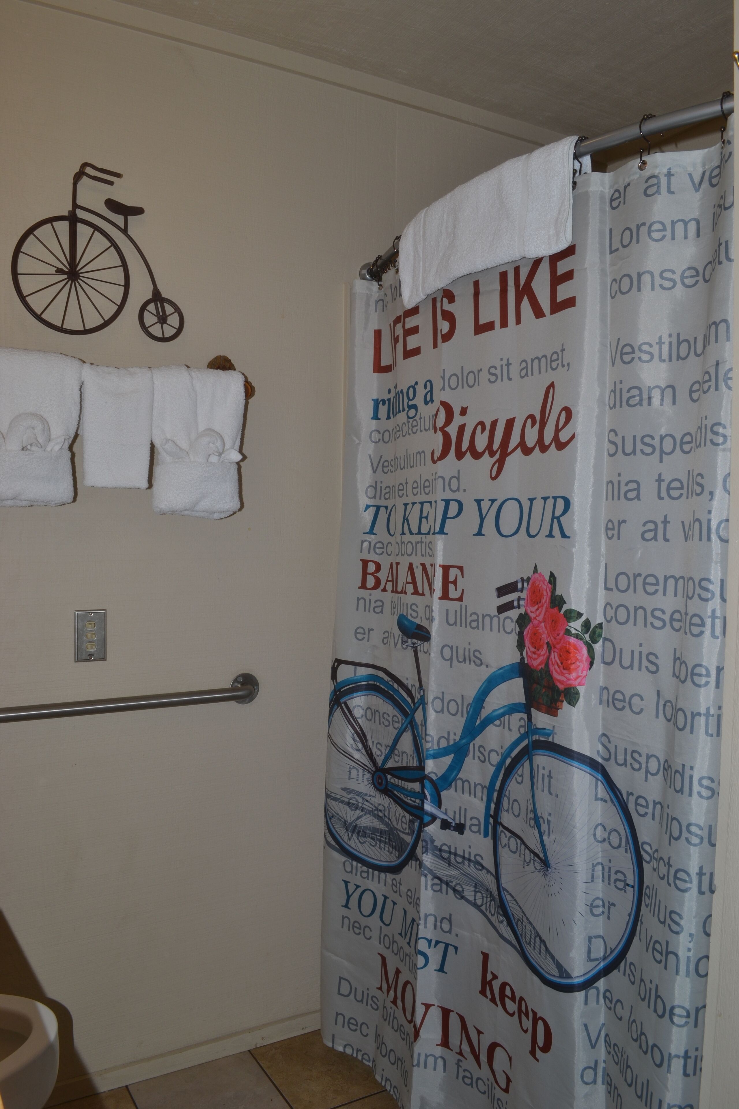 Une salle de bain avec un rideau de douche orné d’un motif de vélo et d’un texte, un porte-serviettes avec des serviettes et une barre d’appui.