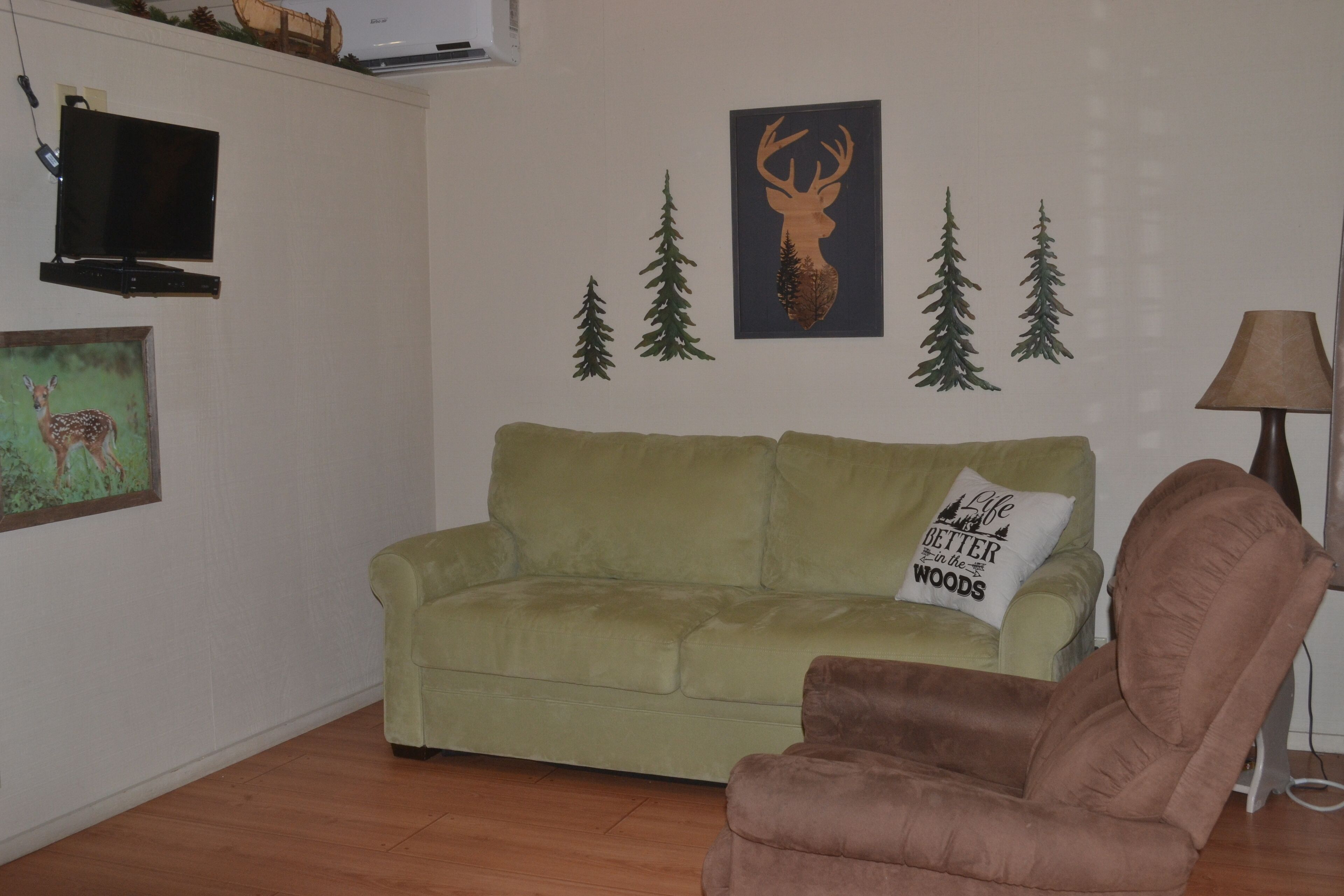 A living room with a green sofa, a brown armchair, a mounted TV, and wall decorations including a deer, a deer head, and trees.