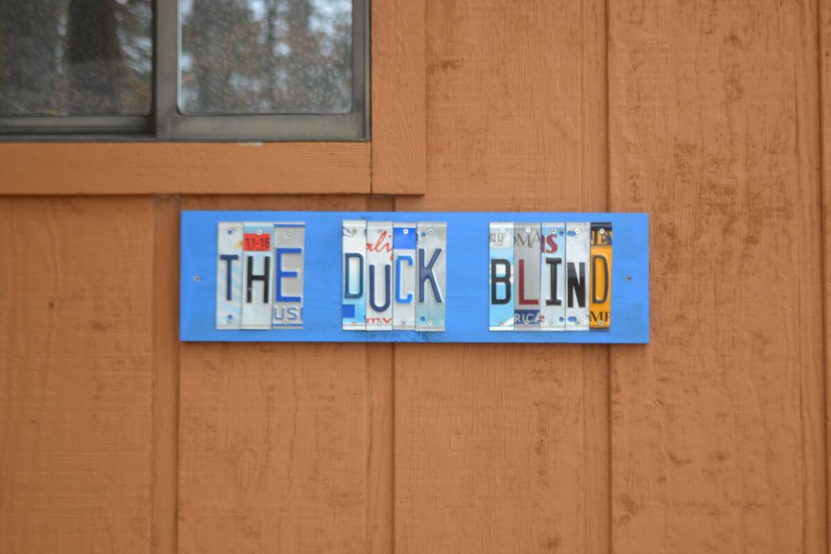 Eine Holzwand mit einem blauen Schild, das Nummernschilder zeigt und den Schriftzug "THE DUCK BLIND" trägt.