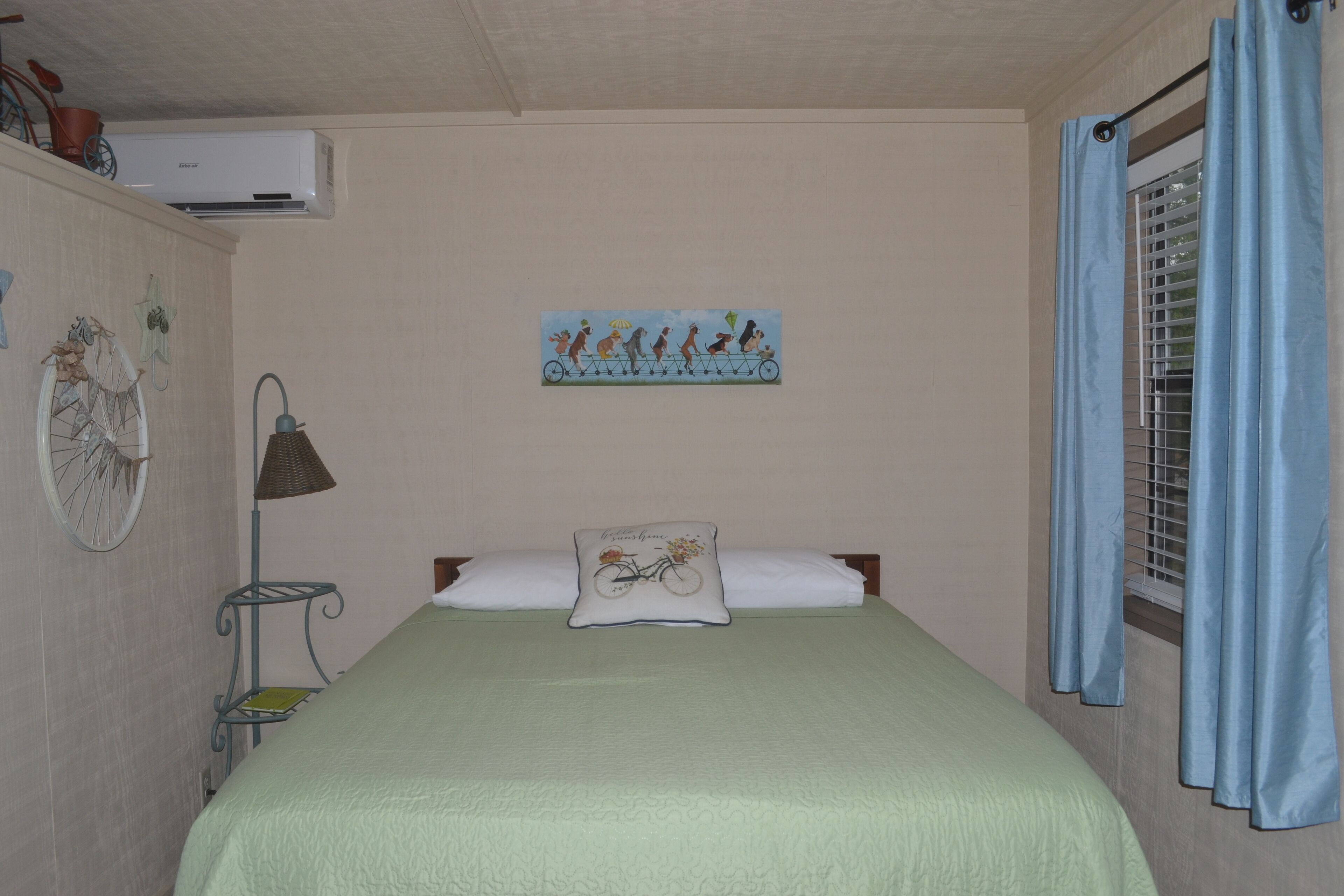 A bedroom with a bed, a wall-mounted air conditioner, a bicycle-themed pillow, a wall decoration, a window with blue curtains, and a small shelf with a lamp.