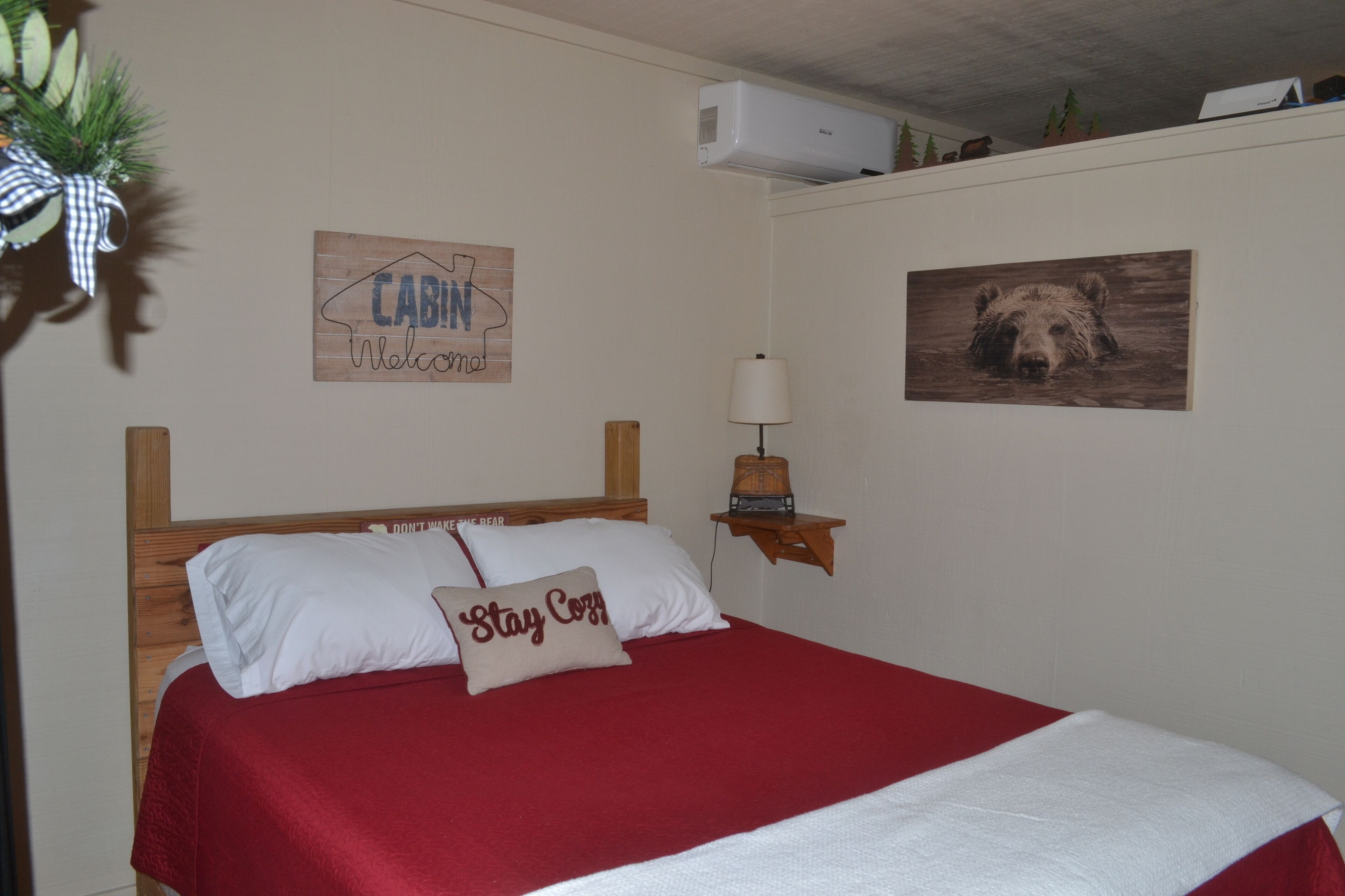 A neatly made bed with a red bedspread and white pillows, a wall-mounted lamp, a welcome sign, and a bear painting.
