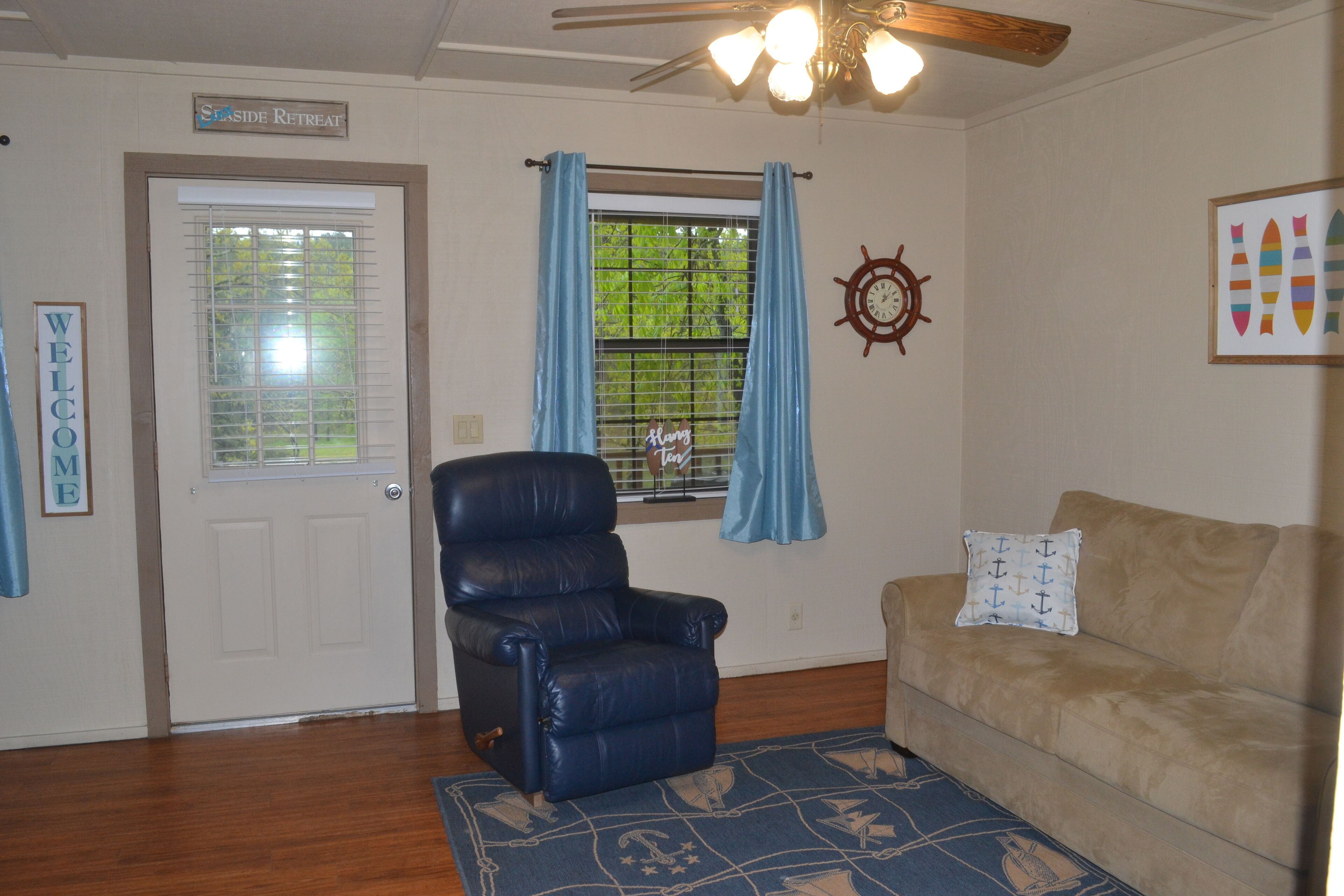 Una sala de estar con un sillón reclinable de cuero, un sofá color beige, un tapete azul, una puerta con un letrero de 'Welcome' y una ventana con cortinas azules.