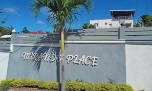 A sign with 'MIRANDO PLACE' written on a grey wall, a palm tree, and a building in the background.