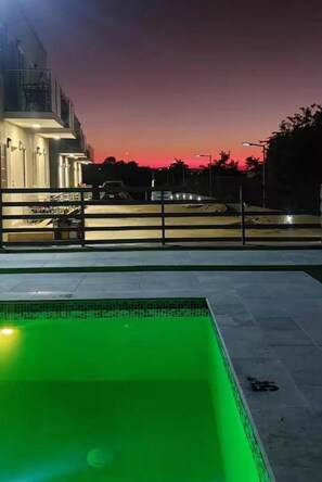 A swimming pool with a green tint, a building with a lit facade, and a sunset sky.
