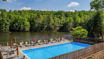 An outdoor swimming pool with lounge chairs and umbrellas, surrounded by a wooden fence and a forested area.