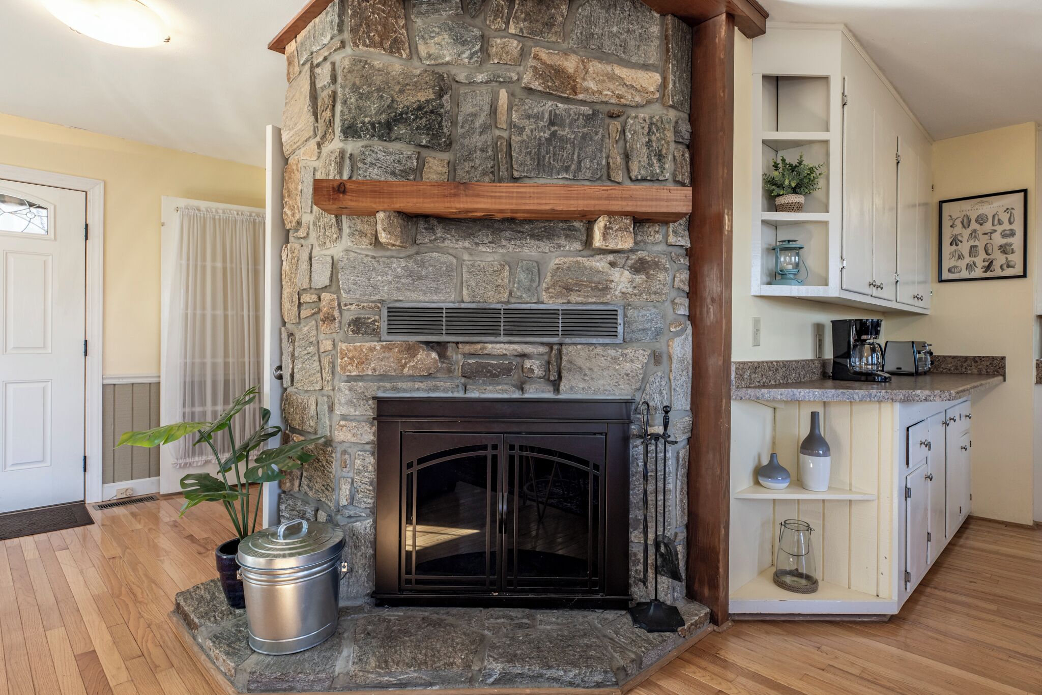 A stone fireplace with a metal vent, a potted plant, and a kitchen area with cabinets and a countertop.