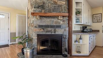 A stone fireplace with a metal vent, a potted plant, and a kitchen area with cabinets and a countertop.