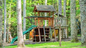 A wooden play structure with a slide and climbing area, surrounded by trees.