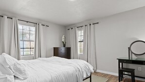 Ein Schlafzimmer mit einem Bett, einem Schminktisch, einem Spiegel und einem Fenster mit Vorhängen.