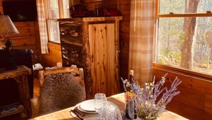 L’intérieur chaleureux d’un chalet avec une table à manger en bois dressée pour un repas, une cheminée et une fenêtre donnant sur la forêt.
