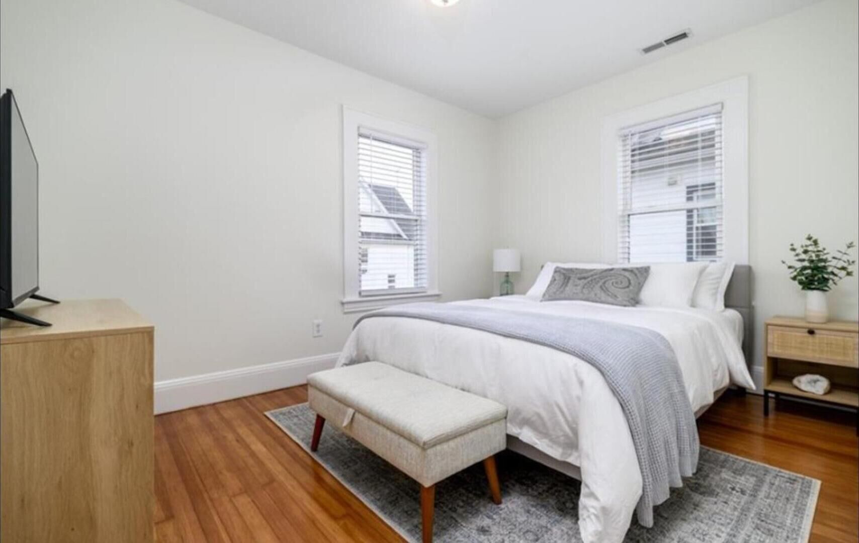 A bedroom with a bed, a bench, a TV on a wooden stand, and two windows with blinds.