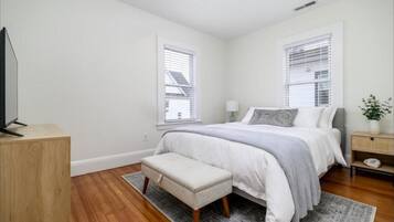 A bedroom with a bed, a bench, a TV on a wooden stand, and two windows with blinds.