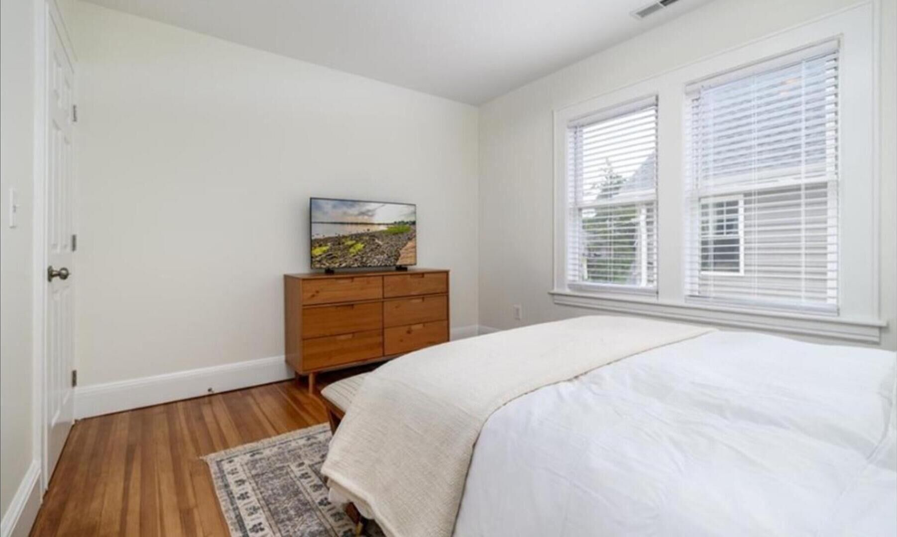 A bedroom with a wooden dresser, a bed with white bedding, a rug, and a window with blinds.