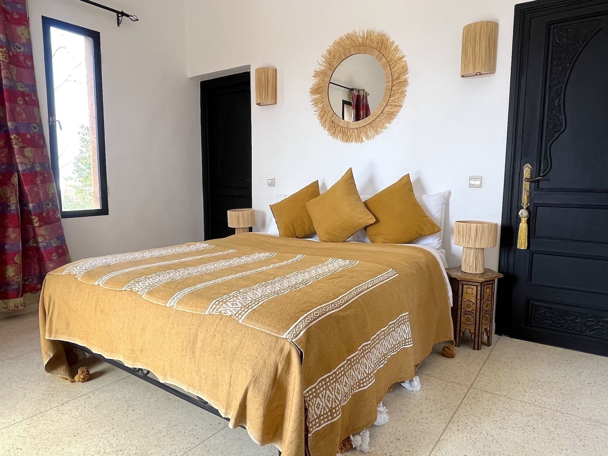 A bedroom with a bed, a mirror, two wall-mounted lamps, a door, and a window with curtains.